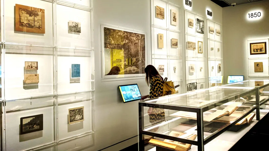 The exhibition wall arranged by decade, with a kiosk between display cases